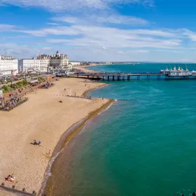 A seafront photo of Eastbourne taken from the west side. The pier can be seen jutting out into the sea towards the top of the photo, as the beach extends round to the left and the emerald sea takes up most of the rest of the shot. The sun is shining and the white hotels on the seafront are very noticeable.