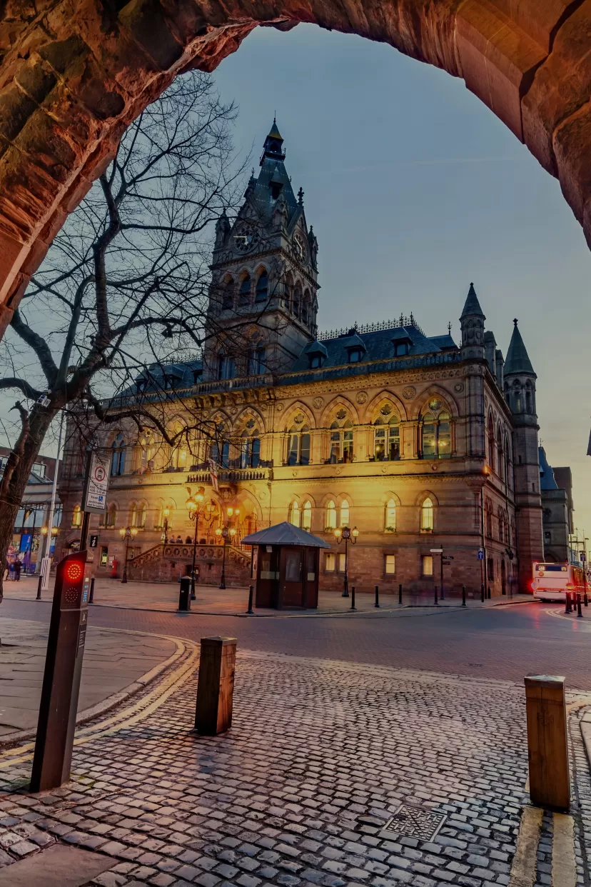 A dusk photo of Chester town hall where the front facade is lit by yellow lighting, whilst the sky is still quite light. The photo is taken from across the square though the Abbey Gateway.