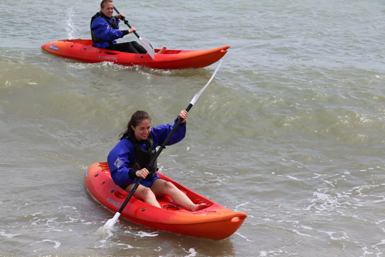 Two of our students kayaking down a wave in Eastbourne. The kayaks are bright red and they are wearing blue windproof tops and life jackets. This is part of the English and watersports option available at ELC Eastbourne in the UK.