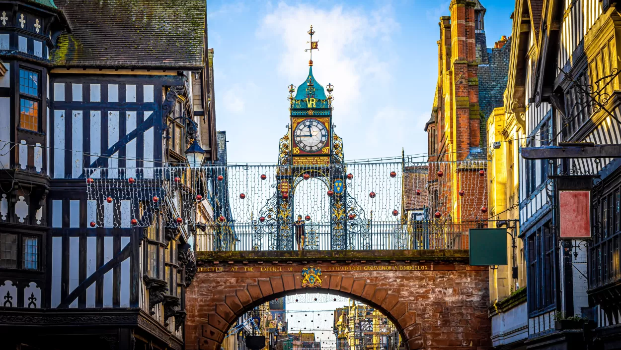 A photograph of the famous Eastgate clock in Chester which stands high above the road on a section of the Roman walls. The clock has a round white face with black roman numerals and hands, and lots of ornate black metalwork filled in with orange and gold panels. The letters VR are above it, visible against the turquoise dome roof, and the date 1897. This was erected in memory of Queen Victoria's diamond jubilee.