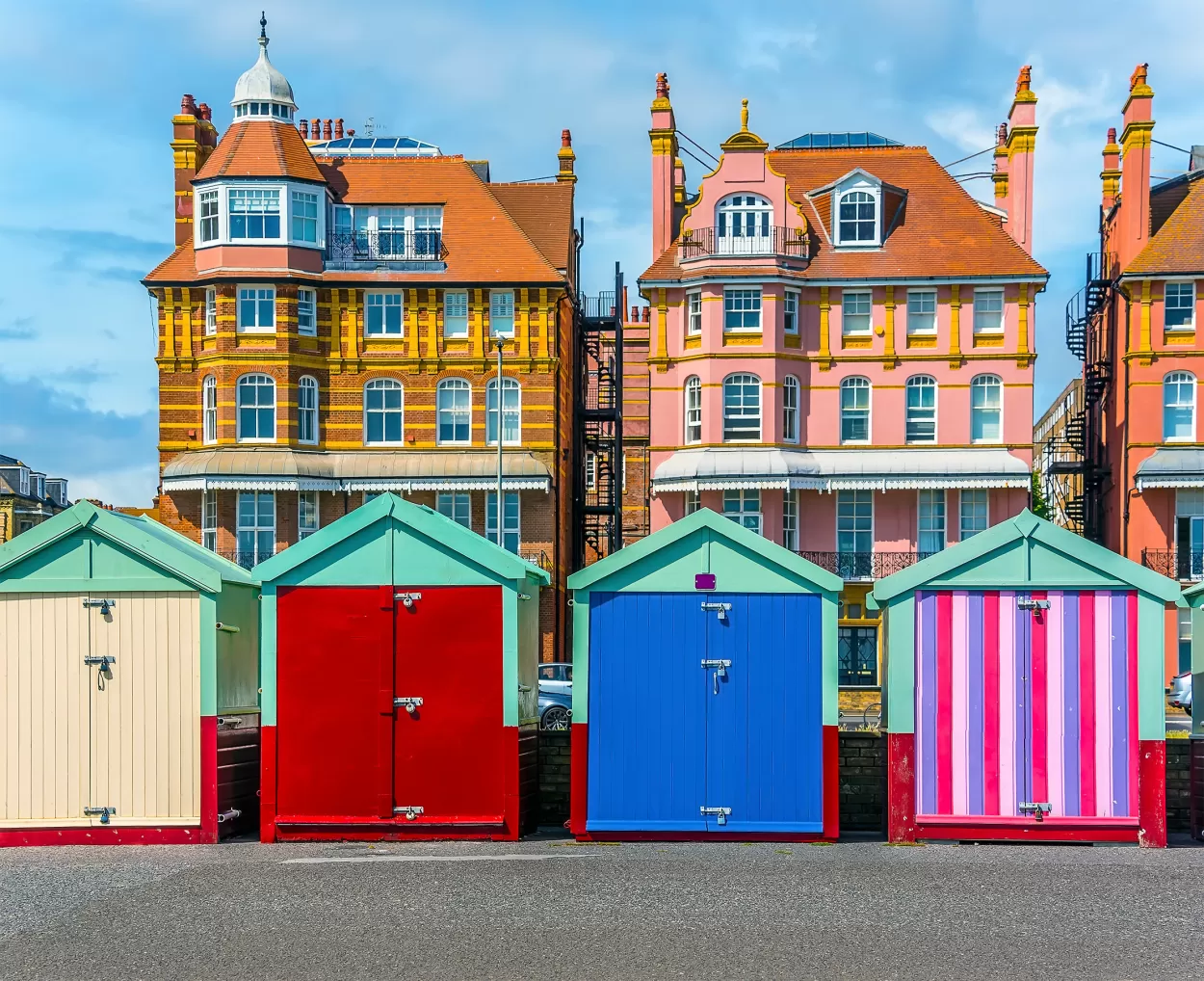 A view of five brightly painted Brighton beach huts which run along the promenade. Behind are three identical looking houses which look very grand, and which, today, will house a number of individual apartments. The sky in the background is very blue.