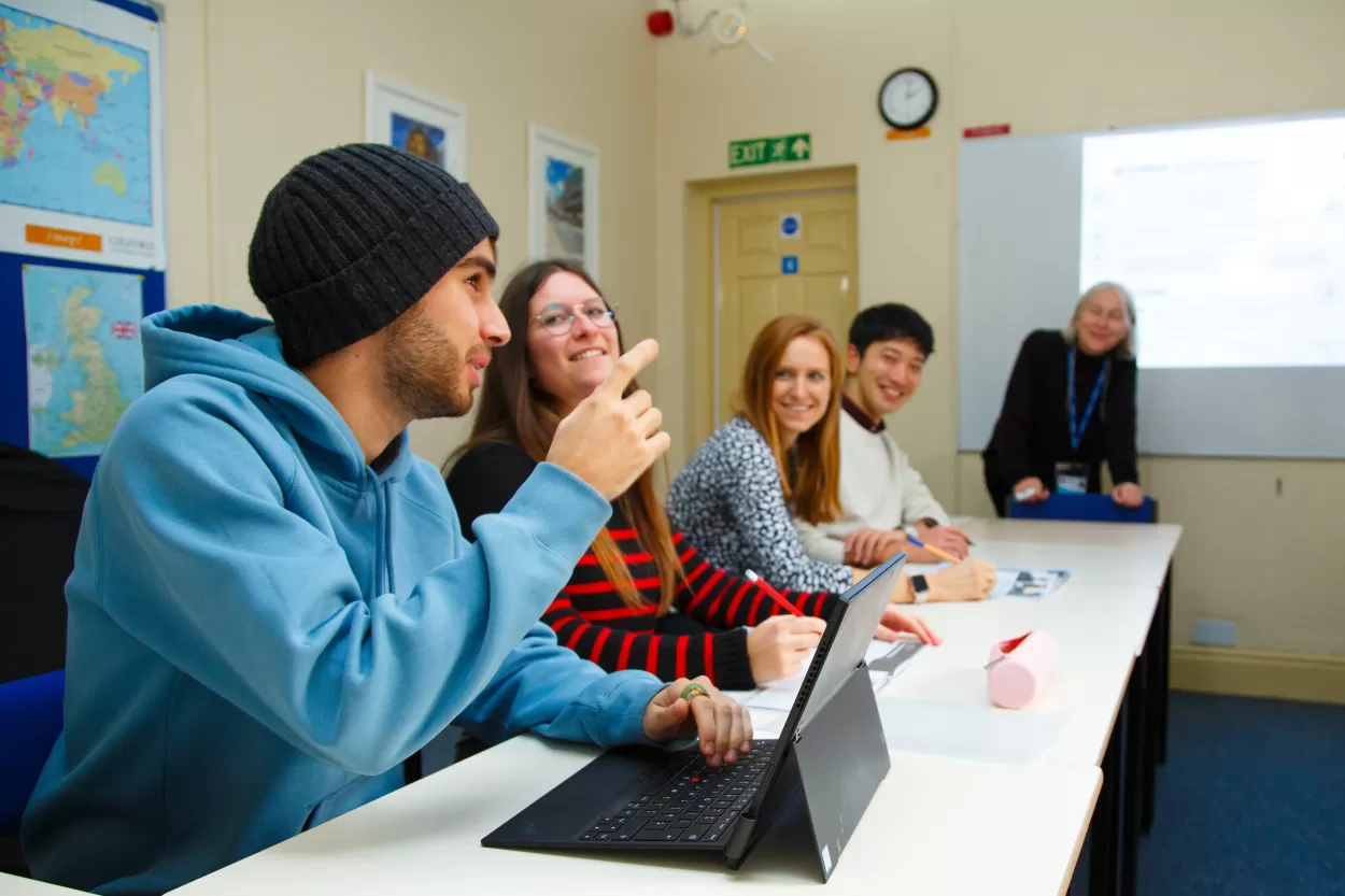 A photo showing four students in class in the English language centre in Chester. The student nearest the camera is wearing a blue hoodie and black beanie and he is making a point and has his forefinger raised emphatically. The other students are smiling and in the background we can see the teacher by the whiteboard looking on.