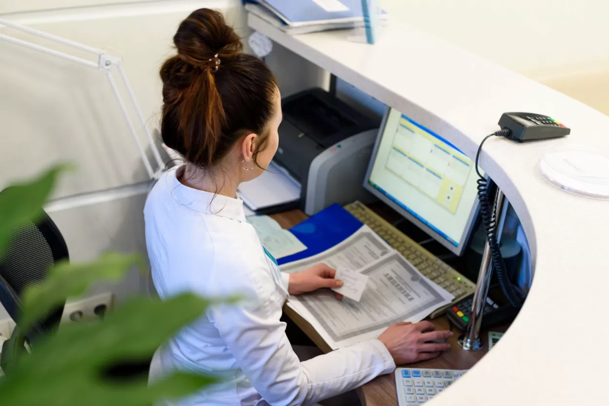 A photograph of an office worker possibly a receptionist sitting at a desk looking at her computer screen which is hidden behind a counter top which is curved and of a white material. There is the terminal of a credit card reader on the desk which further suggests that this person is a receptionist or secretary of some sort. This image is being used to highlight the need for specialist English language skills which ELC offers in its English school in Brighton.