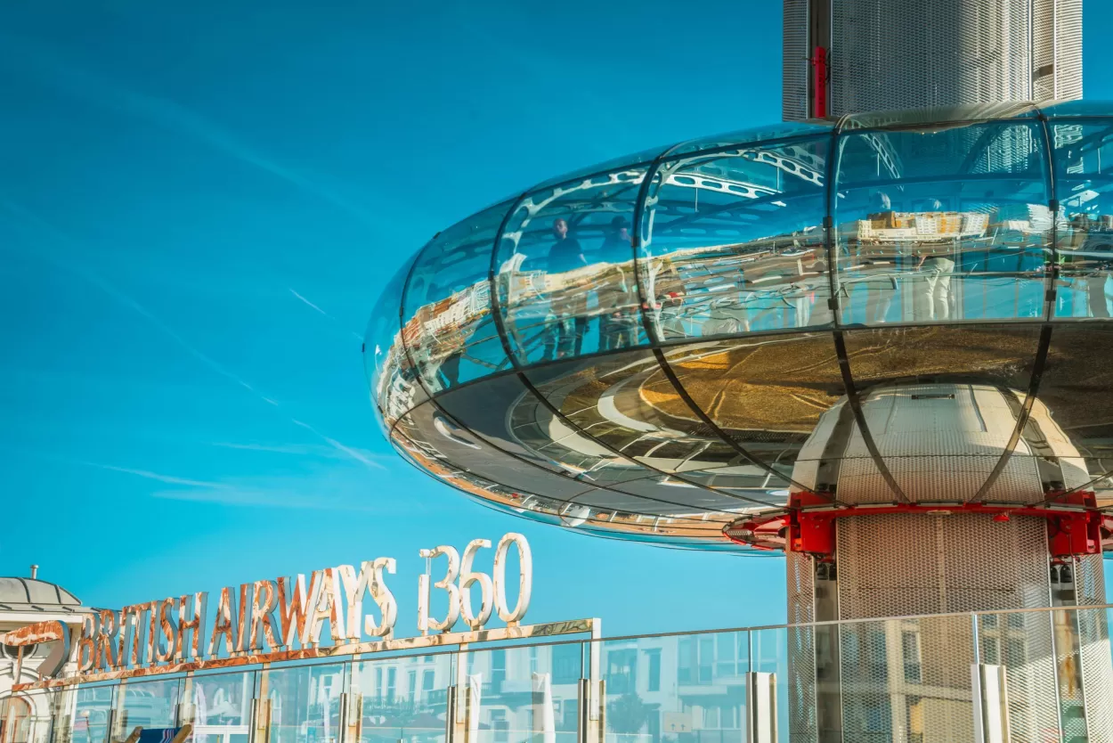 This photo shows the shiny glass, doughnut shaped observation pod of one of Brighton's most recent tourism additions - the i360, which is a 162 metre tall moving observation tower on Brighton seafront. The sky behind this structure is blue and is partly reflected in the glass.