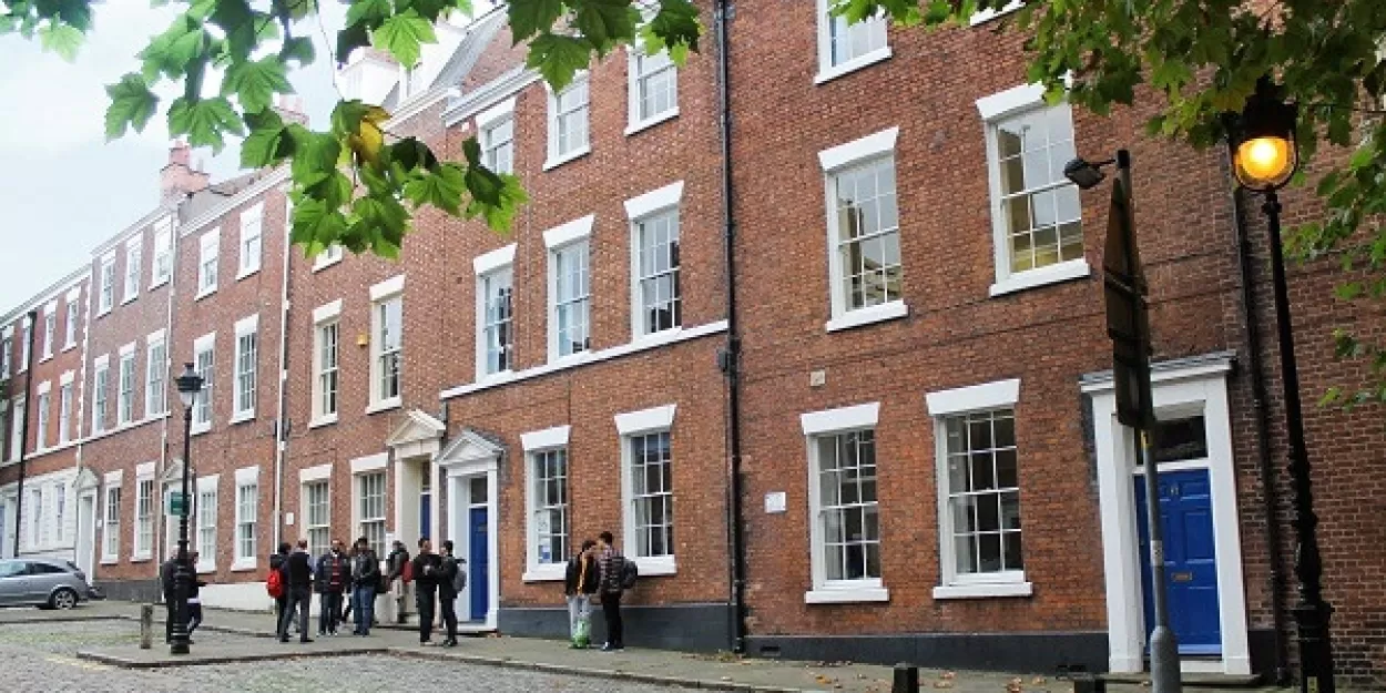 This photo shows the red brick buildings which make up ELC in Chester. The main part of the image shows a three-storey high brick terrace of town houses, with blue doors. The street is cobbled and there is a tree in the foreground whose green leaves overhang the top of the picture.