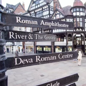 A close-up shot of some of the metal signs in Chester. These are black cast iron signs attached to a post, pointing to different landmarks, labelled in white writing. The signs we can see are to the Roman Amphitheatre, the River & The Groves, Deva Roman Centre. In the background you can see some of the mock-tudor buildings in white and black.