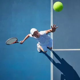 A library photo taken from above of a young man mid-serve during a tennis game. The ball is in the foreground above the player who is poised, racquet behind him. The tennis course is blue and the photo is very striking.