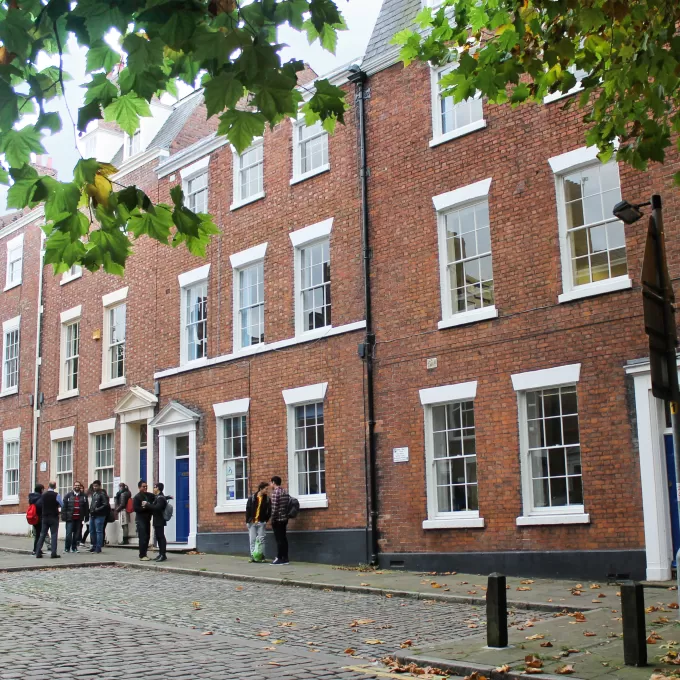 This photo shows the red brick buildings which make up ELC in Chester. The main part of the image shows a three-storey high brick terrace of town houses, with blue doors. The street is cobbled and there is a tree in the foreground whose green leaves overhang the top of the picture.