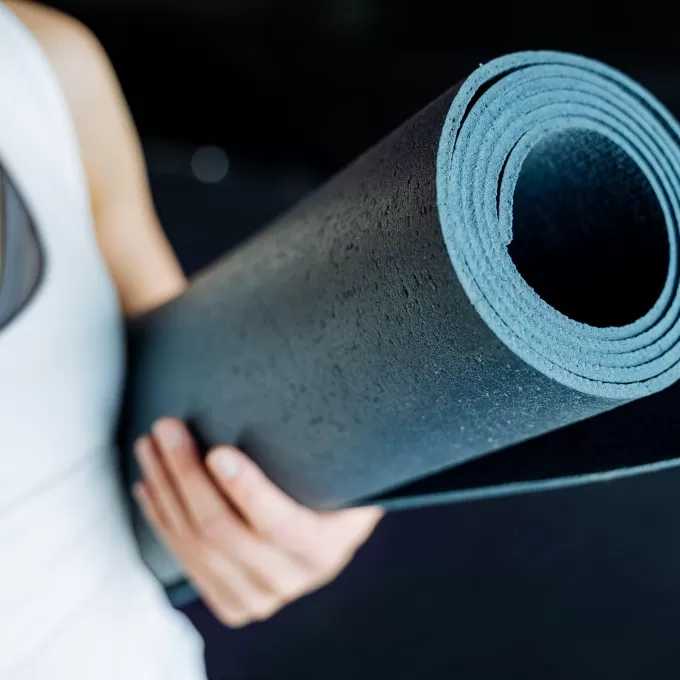 A photo of a rolled up yoga mat in a turquoise-blue colour, being held by someone wearing a white vest top over a dark grey top. The background is blurred. This photo is being used to represent the English plus yoga and natural health class available in Eastbourne, UK