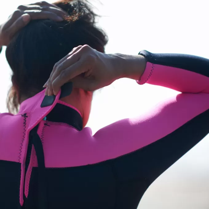 A close up shot of a person fastening the neck flap of a pink and black wetsuit whilst lifting their hair out of the way of the zip and velcro. It looks like they're just getting ready for a watersports activity as part of ELC Eastbourne's English and Watersports course in Eastbourne, UK. The background is completely blurred, but you might think they're looking out to sea.
