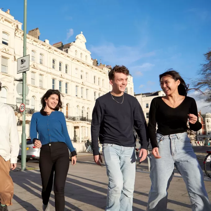 Four smiling language students walk as a group into Palmeira Square in Brighton, with the ELC Brighton school building in its white terrace behind them.