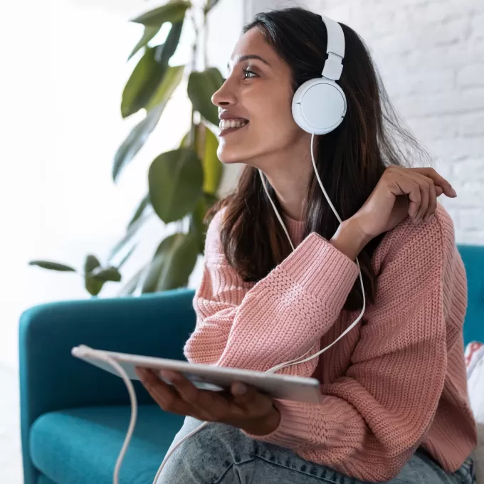 A woman sits on a smart turquoise sofa, with a large indoor plant and whitewashed brick wall in the background. She's listening to something on white headphones connected to her tablet, and she's smiling about it.