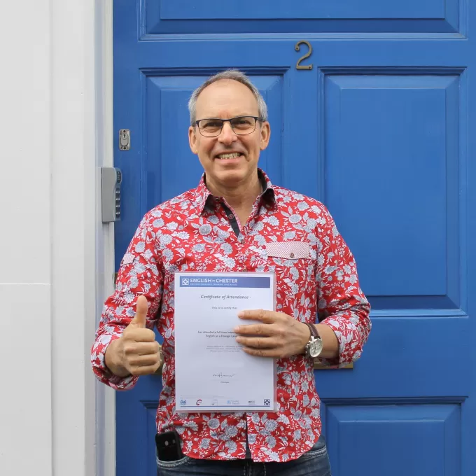 A very happy man in a jazzy red floral shirt gives the thumbs up to the fact he's been given an end of course certificate from the over 25s English course he's attended at ELC Chester. You can spot the familiar bright blue door behind him.