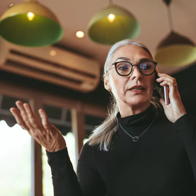 A mature business person talks on her mobile phone whilst holding her other hand up as if to say "well it's obvious" or something like that. She looks slightly concerned. The photograph is taken from down below and behind her head we can see stylish green lamp shades and an air conditioning unit. This is advertising the English courses for executives in Brighton