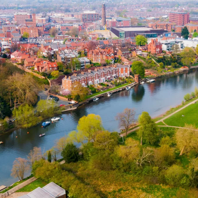 A drone shot of the River Dee in Chester. On one side of the river is the park with lots of different trees, and the other the red brick buildings of all shapes and sizes.