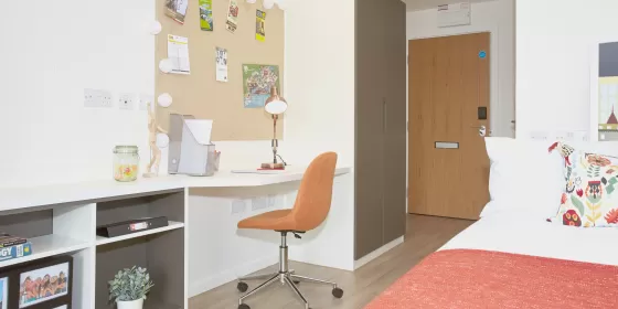 This is a photo of one of the actual ensuite residences used by ELC English language school in Chester. It shows a bed with a salmon pink cover, and similarly toned wheeled office chair at stylish and modern white desk.