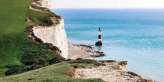 A slightly filtered photo of the the red and white striped Beachy Head lighthouse just outside Eastbourne. You can see the white cliffs and the green grass on top of the cliffs. The sea is a lovely pale blue colour.