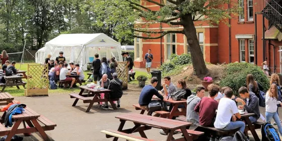 A photo of students sitting at picnic tables chatting together during a break in activities at Loxdale, which is the home to ELC Brighton's summer camp for teenagers. You can see the red brick building in the background and a white gazebo which has been erected on the lawn. The sun is shining.