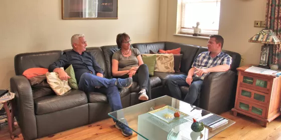 A photo of three adults sitting on a leather couch in the living room of a Brighton home being used for executive students. It appears that the couple hosting are speaking to the student who is wearing jeans and a checked white and blue shirt. The room shows exposed wooden floorboards.