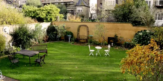 A lovely garden to the rear of the Claremont Hotel in Brighton. You can mainly see a large expanse of grass, and there are a two sets of tables and chairs out. One is wooden, the other is white metal.