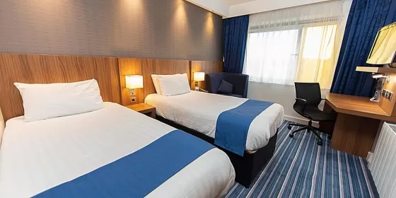 This is one of the photos from the Holiday Inn in Chester, showing two single beds in a modern, blue, white and wood colour coordinated room - there is an office chair and desk in the room. Double beds can also be booked at the hotel.