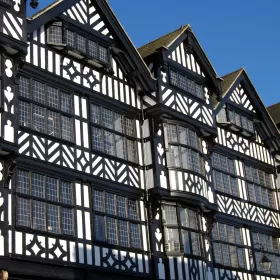 This photo shows some of the shop front above street level in Chester, set against a brilliant blue sky. The buildings have a tudor look to them, built from black timber beams filled with white plaster. The leaded windows are made up of hundreds of small rectangles of glass.
