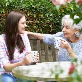 An image of two people sitting outside drinking tea together. They are possible two generations apart, though they are clearly connecting with each other. They're drinking out of identical spotted mugs.