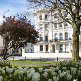 This is a nicely framed image of the front facade of the striking Victorian building which is home to ELC Brighton, one of Brighton's oldest English language schools. In the foreground are white tulips and mature tree and a smaller shrub with red foliage. The school is painted a cream-white colour and the sky is blue with very light white clouds.