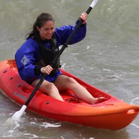 One of our students kayaking down a wave in Eastbourne. The kayak is bright red and she's wearing a blue windproof top and life jacket. This is part of the English and activity programme available at ELC Eastbourne in the UK.