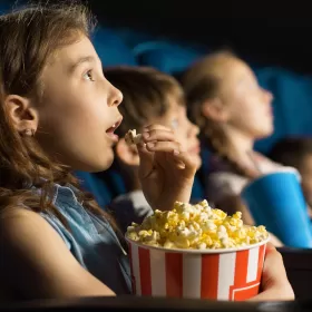 Three quite young children are sitting in a row in a movie theatre. The young girl closest to the camera is staring at the screen looking quite astonished or surprised and she is photographed about to put a piece of popcorn in her mouth and she's holding a large bucket of popcorn in her lap.