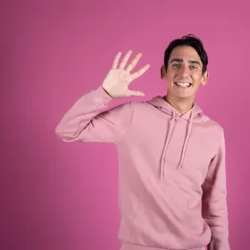 A young man in a pink hoody pictured on a darker pink background. He's smiling and waving in a way that suggests he's trying to attract your attention. This is to represent communication in English and we're thinking about the IELTS speaking test.