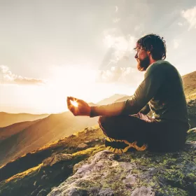 A stylised photo of a man with a beard meditating on top of a mountain. The sun is strategically placed to be shining through the gap between his fingers and his palms. It looks like someone is getting into the right mental space.