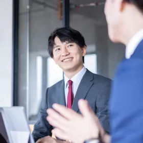 This image shows two men in business suits in an office environment. The one facing us has a dark hair, and a red tie. He's smiling. We're using this to denote negotiation. Maybe they've just reached the end of a particularly tough negotiation in English.
