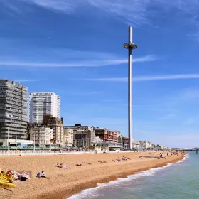This photo shows one of Brighton's most recent tourism additions - the i360, which is a 162 metre tall moving observation tower on Brighton seafront. It looks like a large nail that has been hammered into the beach. The rest of the photo shows the pebble beach, the green sea and a blue sky.