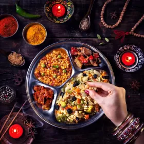 A photo what looks like some very tasty south Asian food, served in a round stainless steel dish. Their are pots of spices and candles dotted about to confirm the origin of the food.