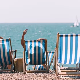 A shot of Brighton's seafront in which the focus is three deckchairs placed so as to look out to see. The fabric of the deck chairs is blue and white stripes. Two of the chairs are occupied with sunbathers. Out to sea a white sailing boat glides out of shot.